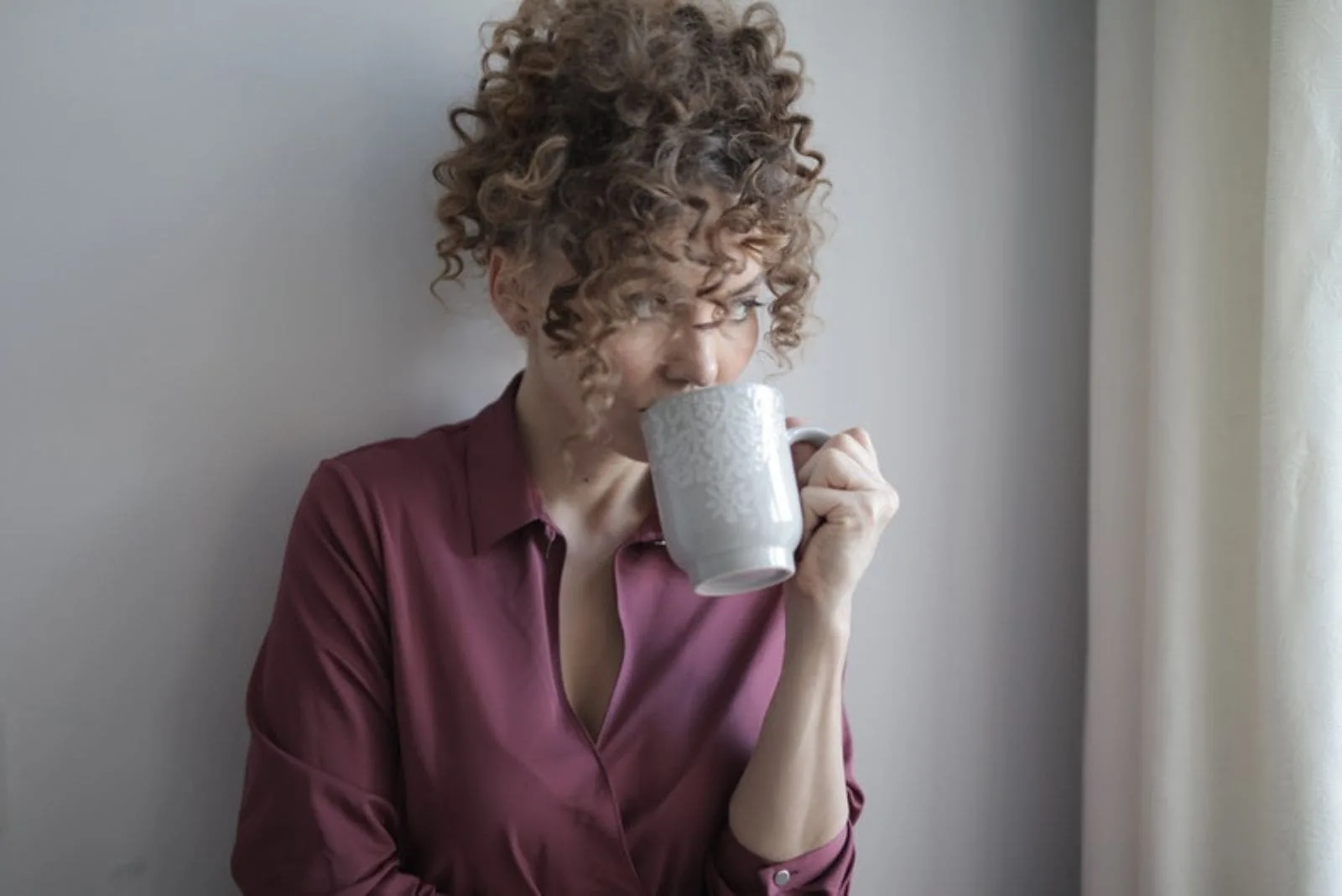 adult curly haired woman leaning on the wall while drinking from a mug looking away out the window