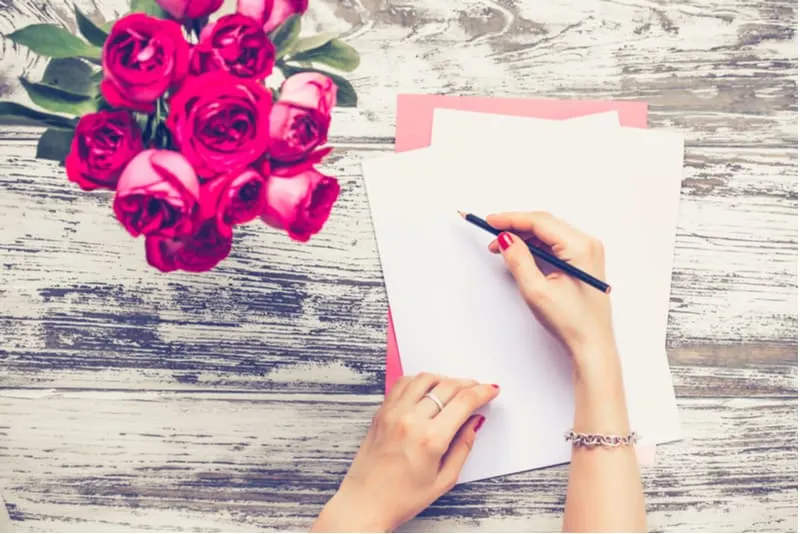 woman's hand writing a letter on the table with vase of red roses